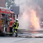 Ein brennendes Auto am Straßenrand, Feuerwehr im Einsatz, öffentlicher Verkehr gesperrt.