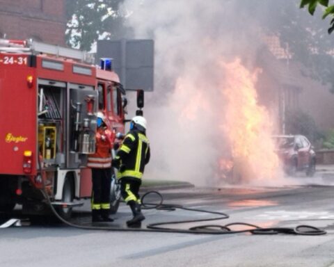 Ein brennendes Auto am Straßenrand, Feuerwehr im Einsatz, öffentlicher Verkehr gesperrt.