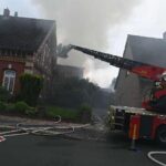 Großer, zerstörter Arbeitsschuppen mit Rauch und Feuerwehrfahrzeugen im Vordergrund. Brandgeruch in der Luft.