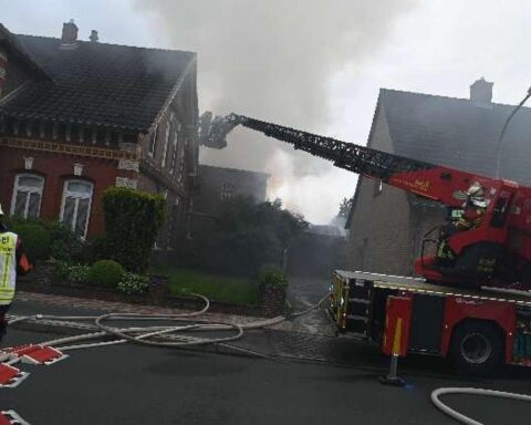 Großer, zerstörter Arbeitsschuppen mit Rauch und Feuerwehrfahrzeugen im Vordergrund. Brandgeruch in der Luft.
