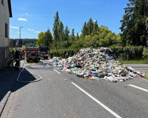 Brennender Müllcontainer auf Straße; Feuerwehr löscht Feuer, Fahrer verhindert Fahrzeugschaden.