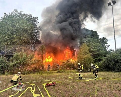 Brennendes Vereinsheim mit dramatischen Flammen und Feuerwehr im Einsatz bei Nacht.