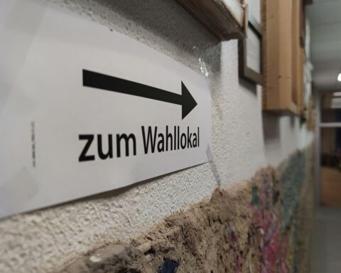 Wahllokal mit Wählern, umgeben von Wahlzetteln und Informationsmaterial, symbolisiert demokratische Mitbestimmung.