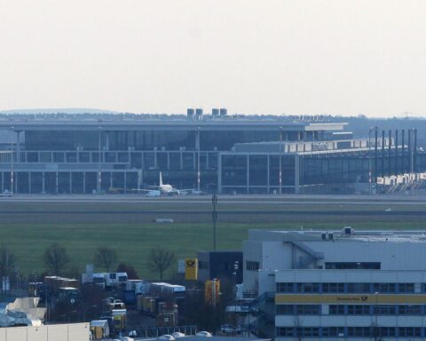 Bild von einem geschäftigen Flughafen mit Passagieren, Gepäck und Flugzeugen im Hintergrund.