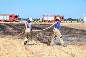 Feuerwehrleute löschen Feldbrand, Rauchentwicklung sichtbar, Einsatzkräfte und Landwirte in Aktion.