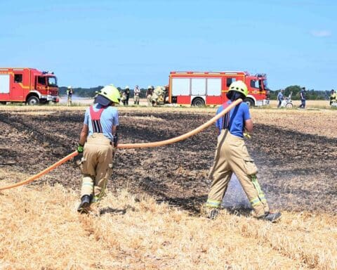 Feuerwehrleute löschen Feldbrand, Rauchentwicklung sichtbar, Einsatzkräfte und Landwirte in Aktion.