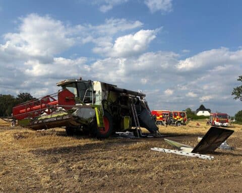 Ausgebrannter Mähdrescher vor einem getoasten Kornfeld, Feuerwehrleute bei Löscharbeiten.