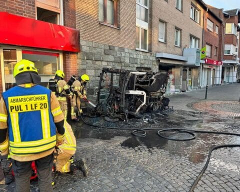 Eine brennende Straßenkehrmaschine in der Fußgängerzone, Einsatzkräfte der Feuerwehr löschen das Feuer.
