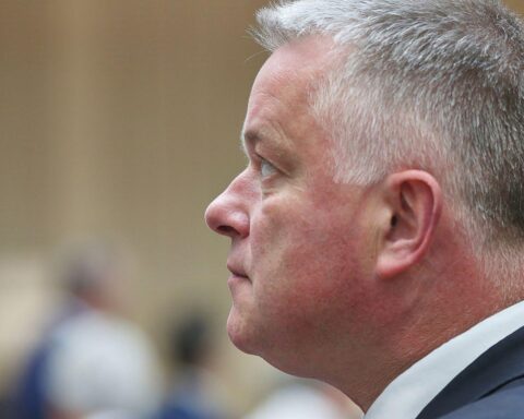 Monika Helfer (Archivfoto) vor Publikum bei Auszeichnung; Porträt des Ministerpräsidenten Gordon Schnieder im Hintergrund.